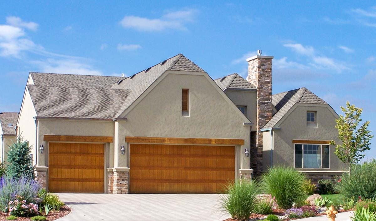 House plan exterior: stucco finish, two-car garage with wood doors, stone accents, and a prominent stone chimney.