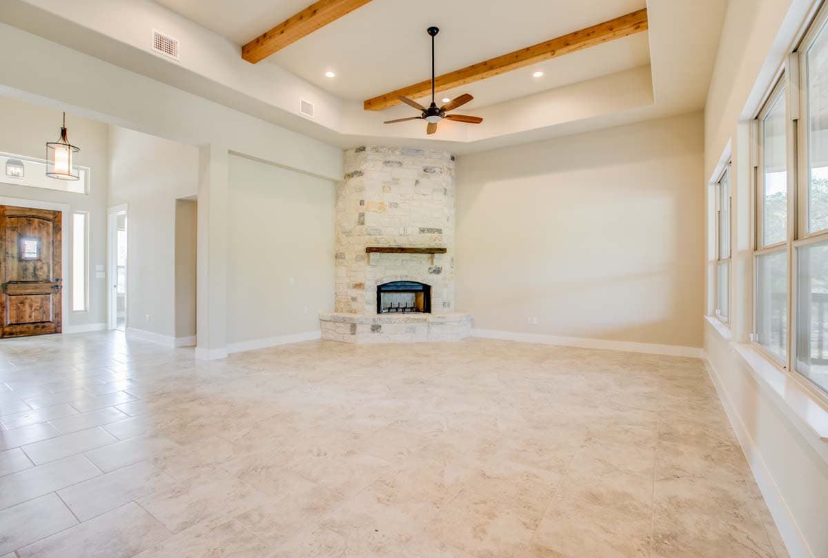 Great room with stone fireplace, tray ceiling with wood beams, ceiling fan, and large windows.