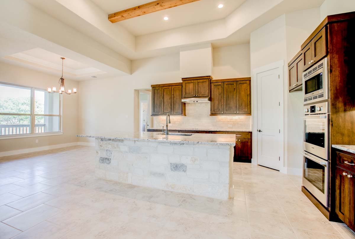 Kitchen with stone island, dark wood cabinets, double ovens, tray ceiling with wood beam, and large windows.