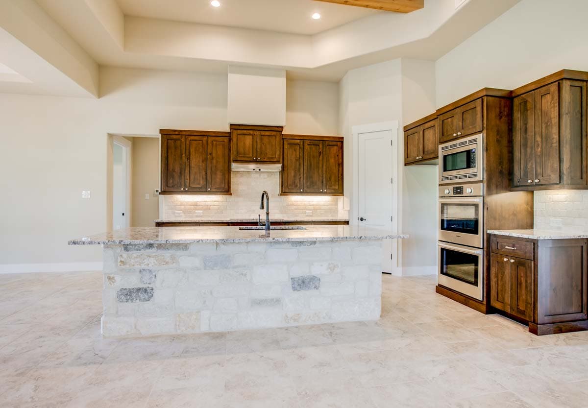 Kitchen with large stone island, dark wood cabinets, stainless steel double ovens, and exposed wood beam ceiling.