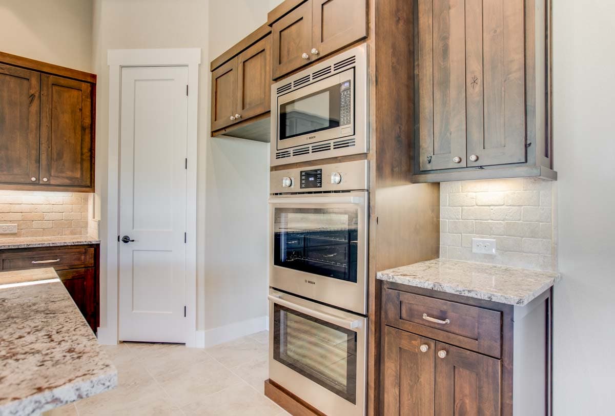 Kitchen with stainless steel double ovens, wood cabinetry, granite countertops, and subway tile backsplash.