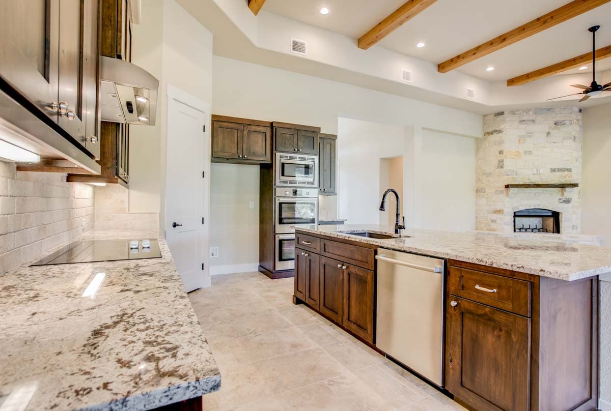 Interior view of kitchen with dark wood cabinets, granite countertops, island, stainless steel appliances, and stone fireplace with exposed beams.