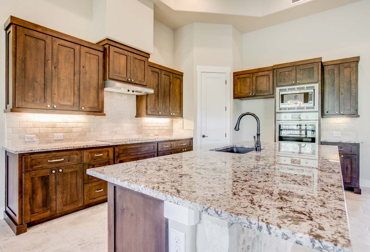 Kitchen with dark wood cabinets, granite countertops, island with sink, subway tile backsplash, and built-in oven and microwave.