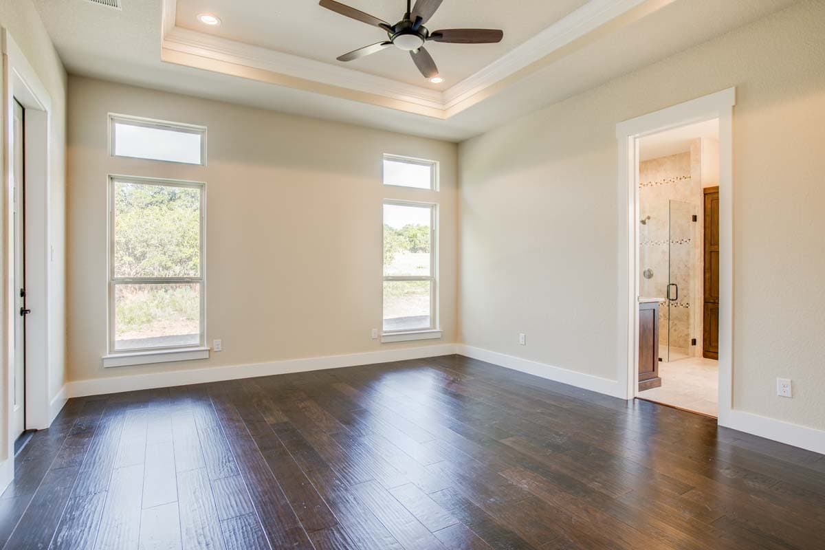 Interior view of a bedroom with dark wood floors, a tray ceiling with fan, and windows overlooking greenery. Ensuite bathroom visible.