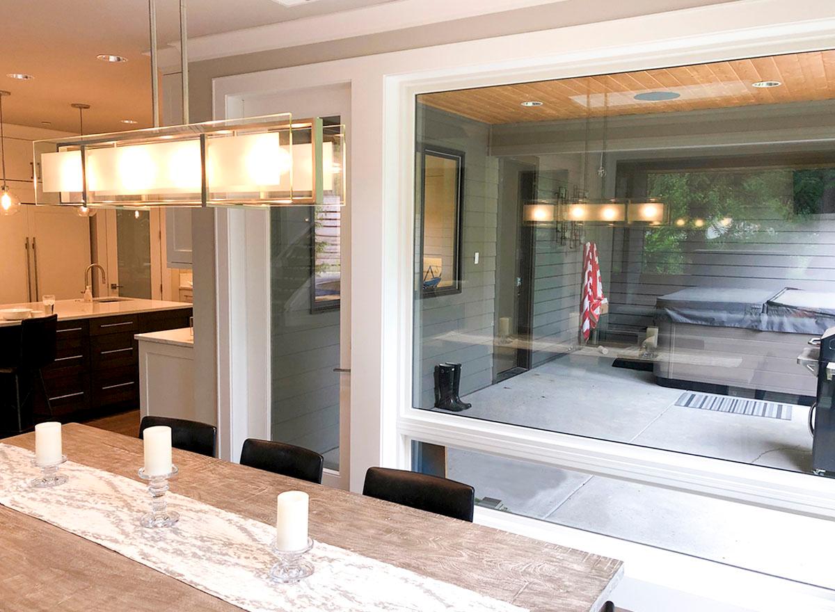 Interior view showing dining area with modern chandelier, kitchen island, and large window overlooking patio with hot tub.