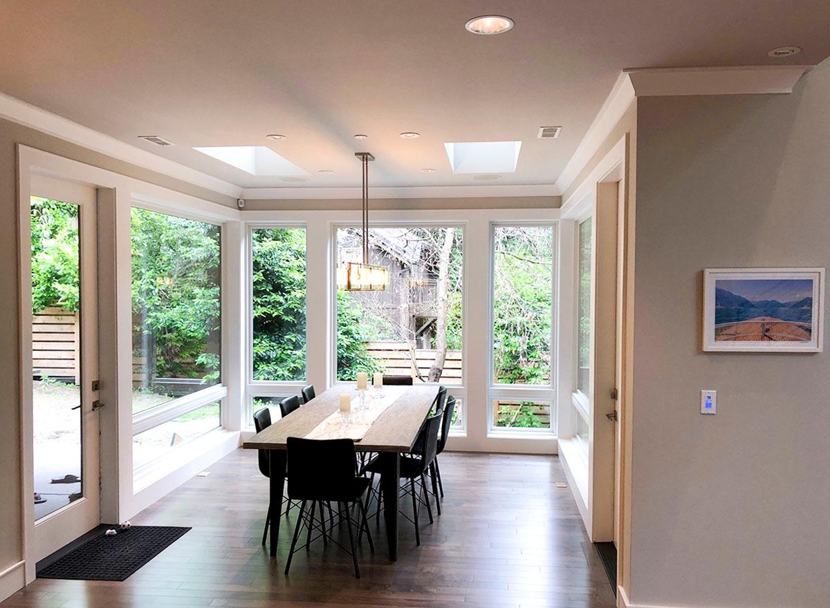 Dining room with large windows, skylights, and a rectangular chandelier above a wood table set for eight.