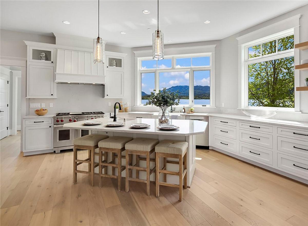 Kitchen interior with white cabinets, a central island with seating, range, and large windows with water and mountain views.