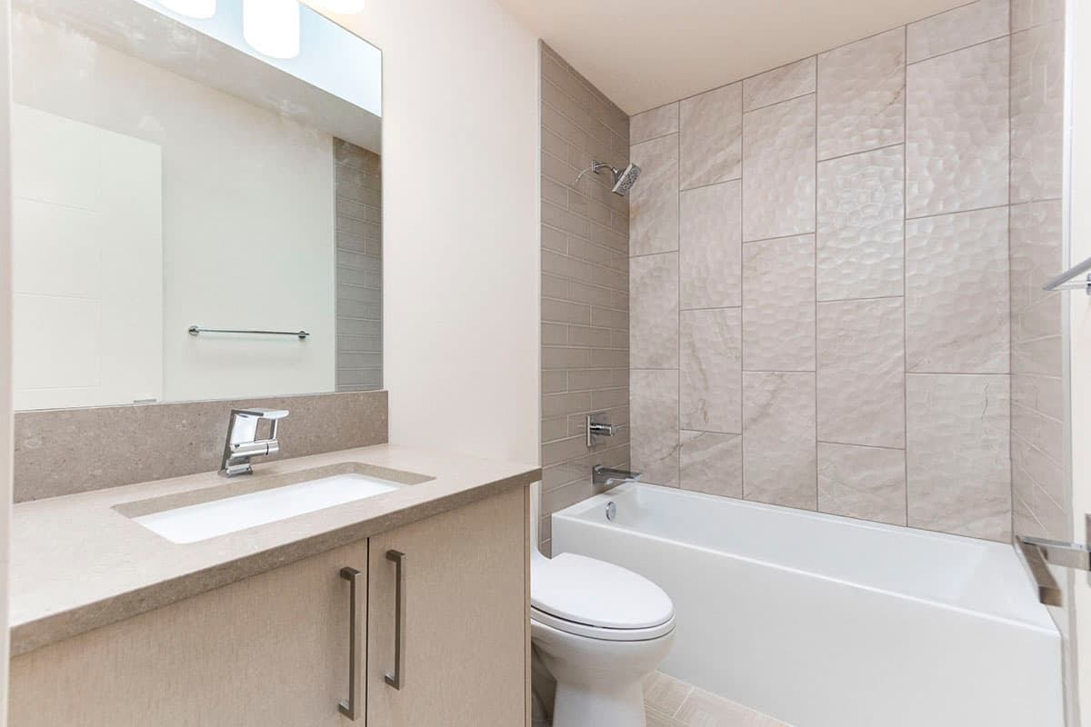 Modern bathroom with vanity, toilet, and bathtub featuring textured tile shower surround and beige wood cabinetry.