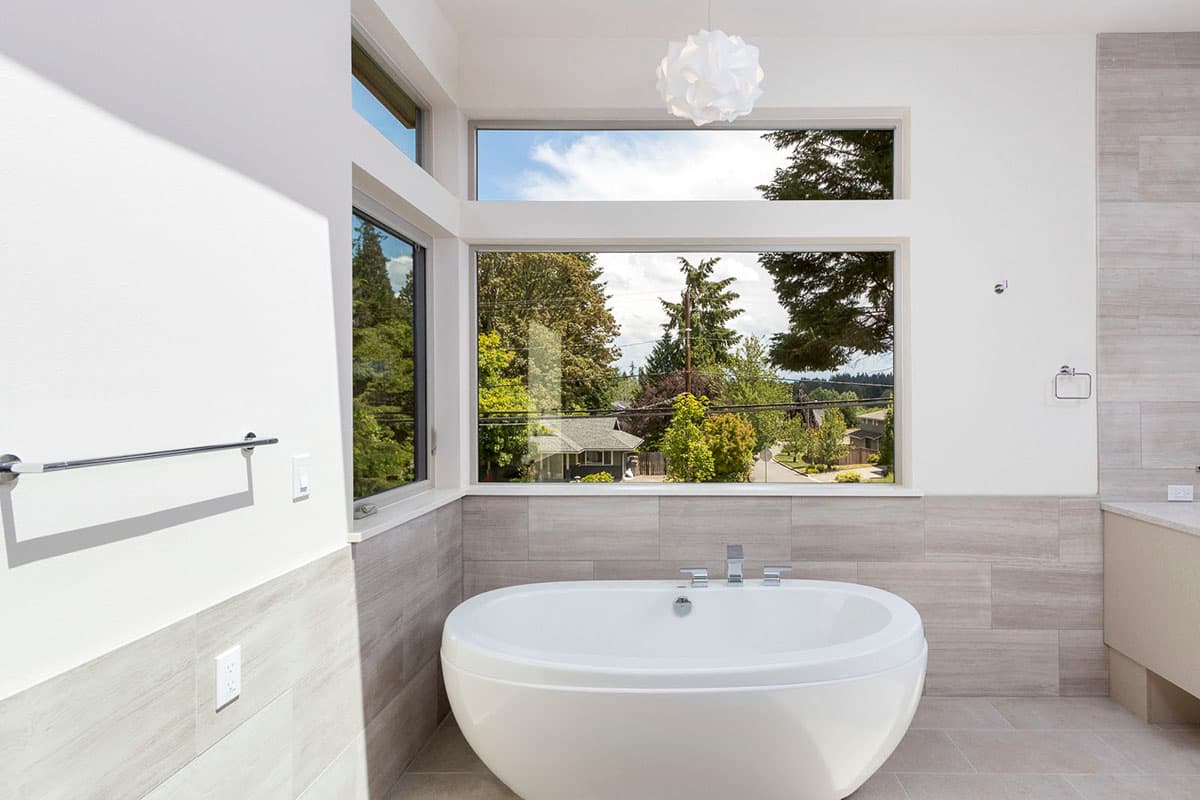 Modern bathroom with freestanding oval tub and large corner windows offering panoramic views of trees and neighborhood.
