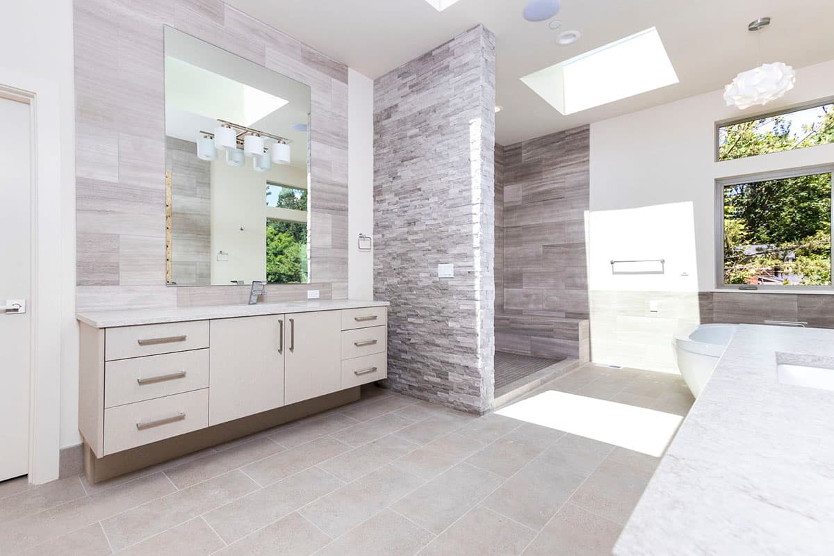 Modern bathroom with double vanity, stone accent wall, walk-in shower with skylight, and large windows.