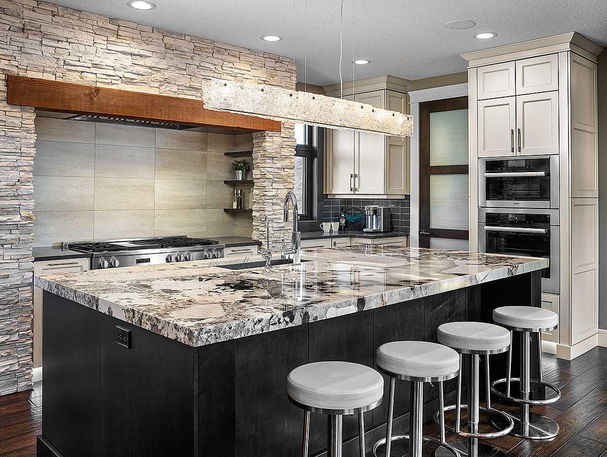 Kitchen interior with a large island, stone accent wall behind the range, and stacked ovens. A modern chandelier hangs above the island.