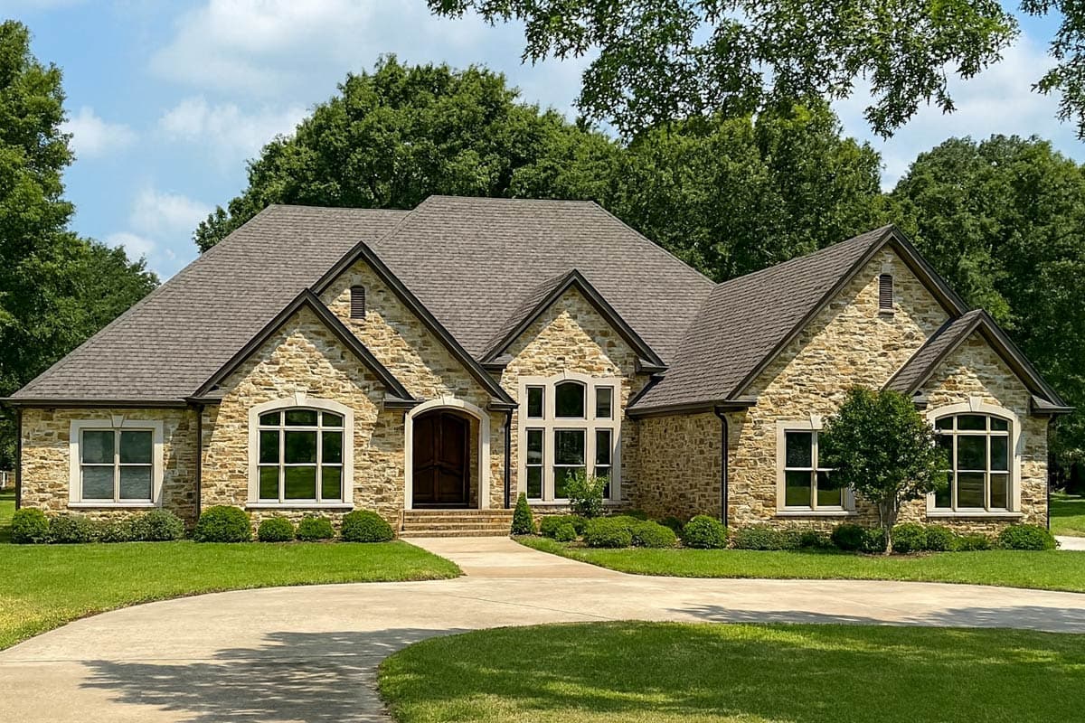 House plan exterior. Stone-clad, single-story home with multiple gabled roof sections, arched entry door, and large multi-pane windows.
