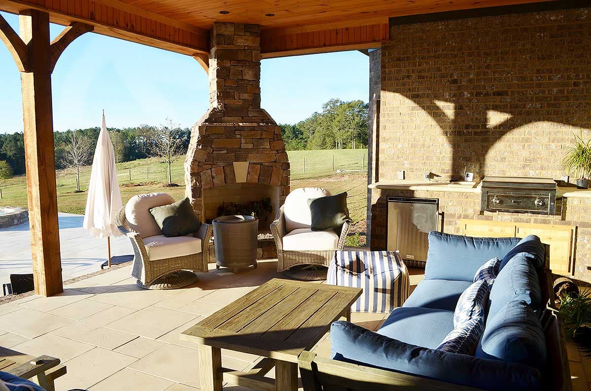 Covered outdoor living space with stone fireplace, seating, grilling station, and wood-paneled ceiling.