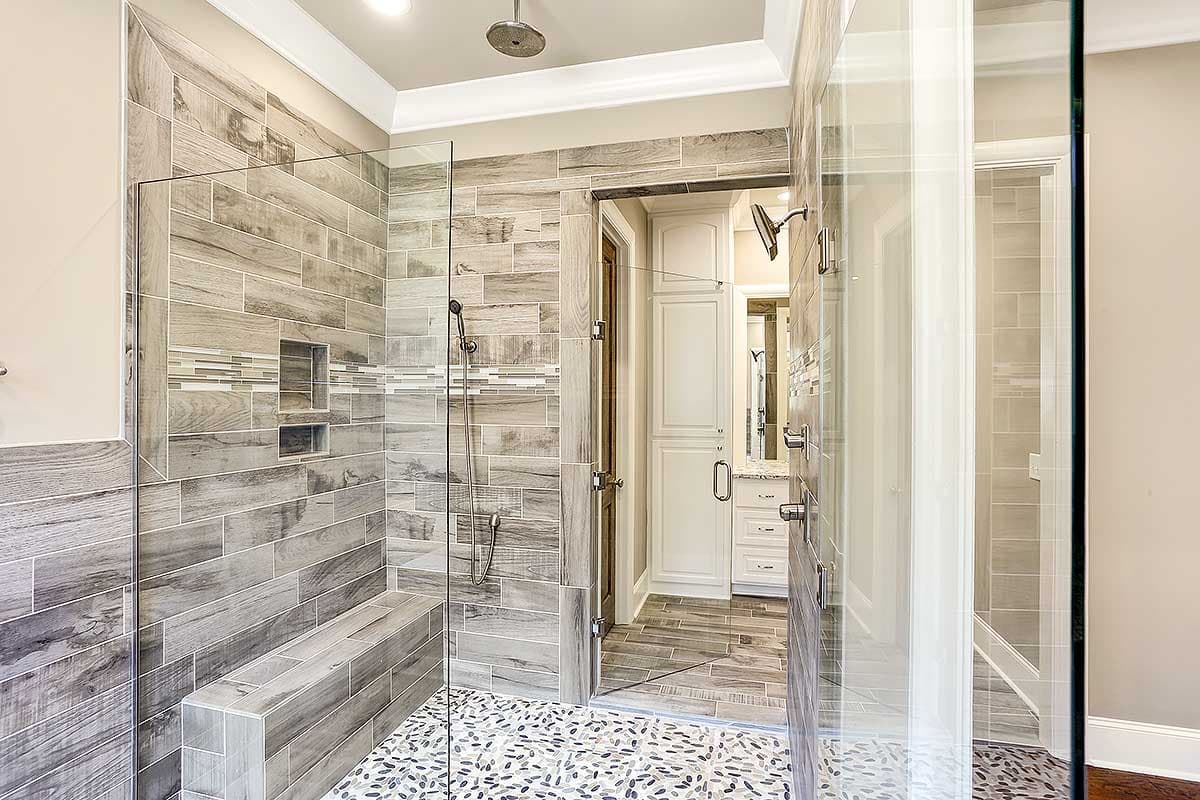 Enclosed glass shower with wood-look tile, built-in bench, and pebble floor, opening to vanity area with white cabinetry.