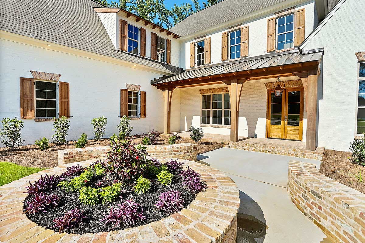 House plan exterior with white brick, wood shutters, covered entry with timber supports, and circular garden beds.