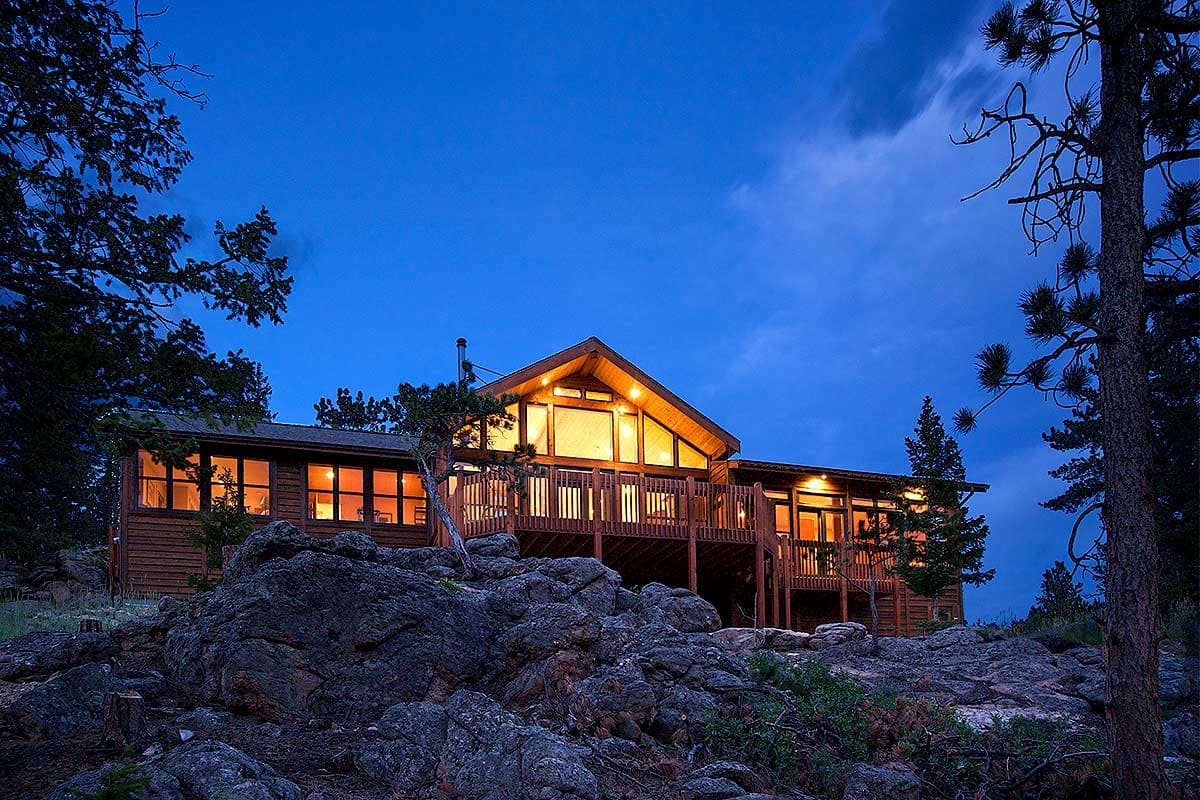 House plan exterior. Two-story home with a gabled roof, large windows, and a wraparound deck. Situated on a rocky hillside.