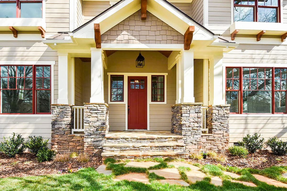 Craftsman style house plan exterior with stone-clad porch columns, a gabled entryway, and red-framed windows.