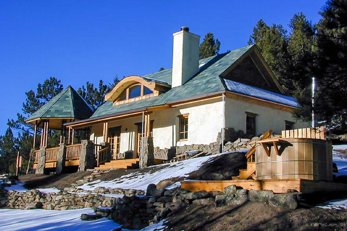House plan exterior with arched dormer, covered porch, stone foundation, and wood hot tub.