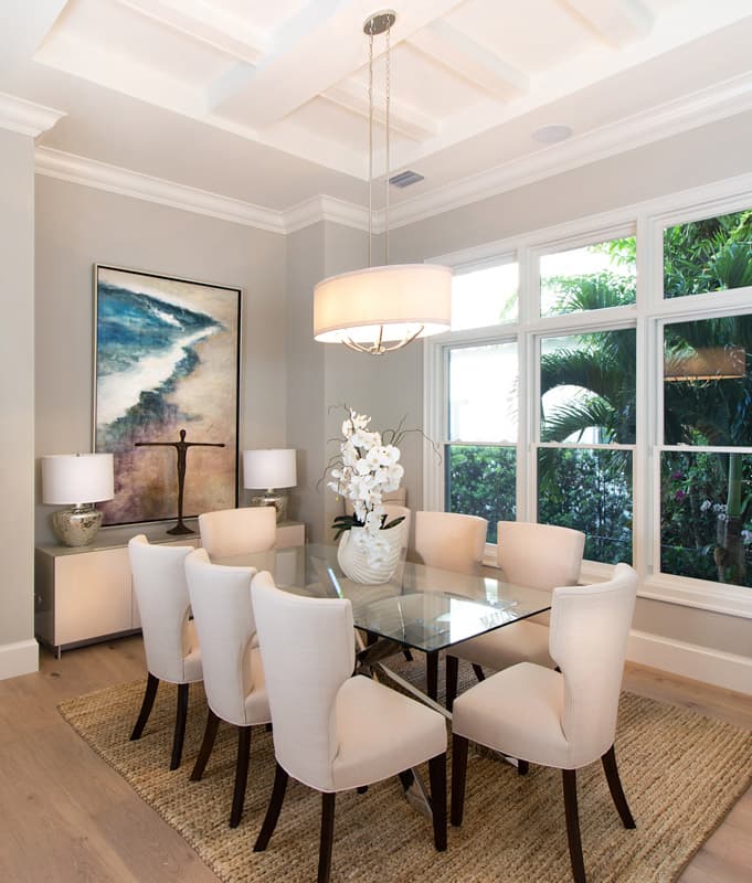 Dining room with glass table, upholstered chairs, coffered ceiling, and large windows with tropical foliage views.