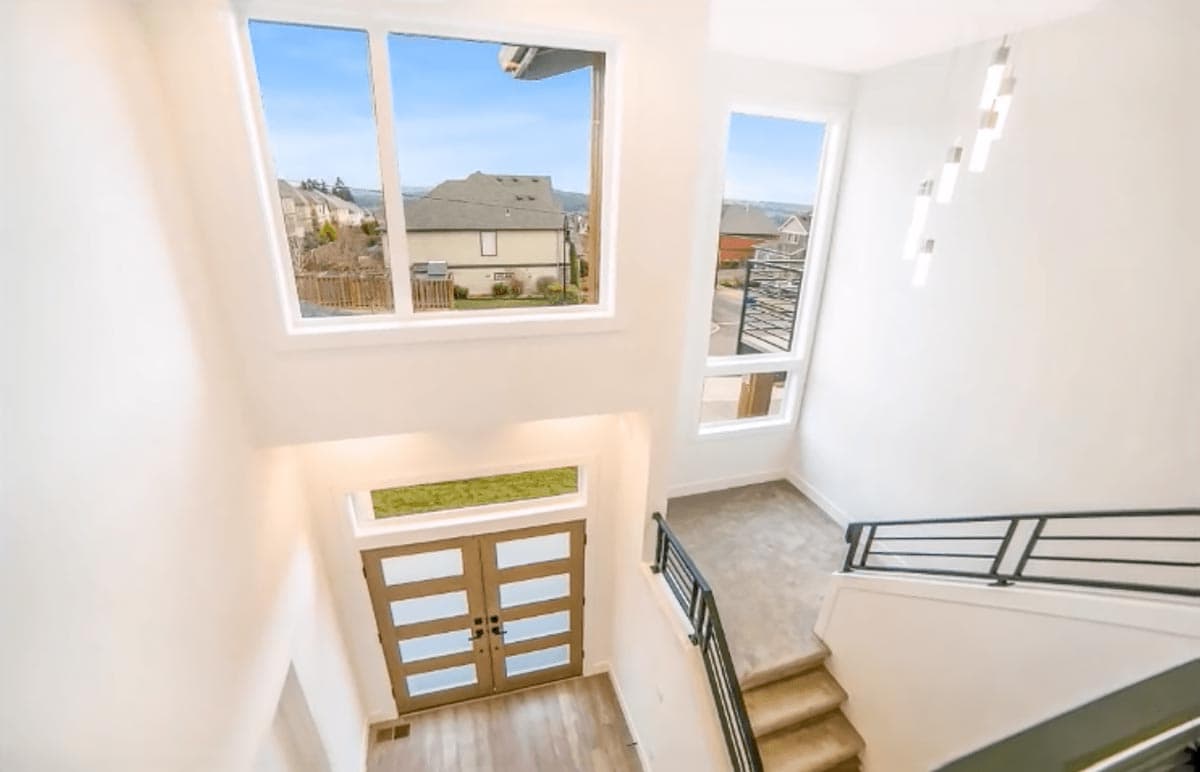 Modern house interior: double glass doors, tall windows, and a staircase with black railing.