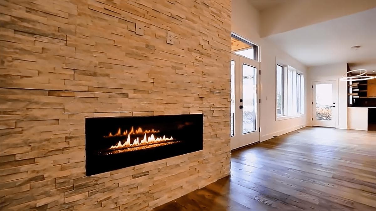Interior view of a modern home featuring a stacked stone fireplace, large windows, and an open layout leading to a kitchen.