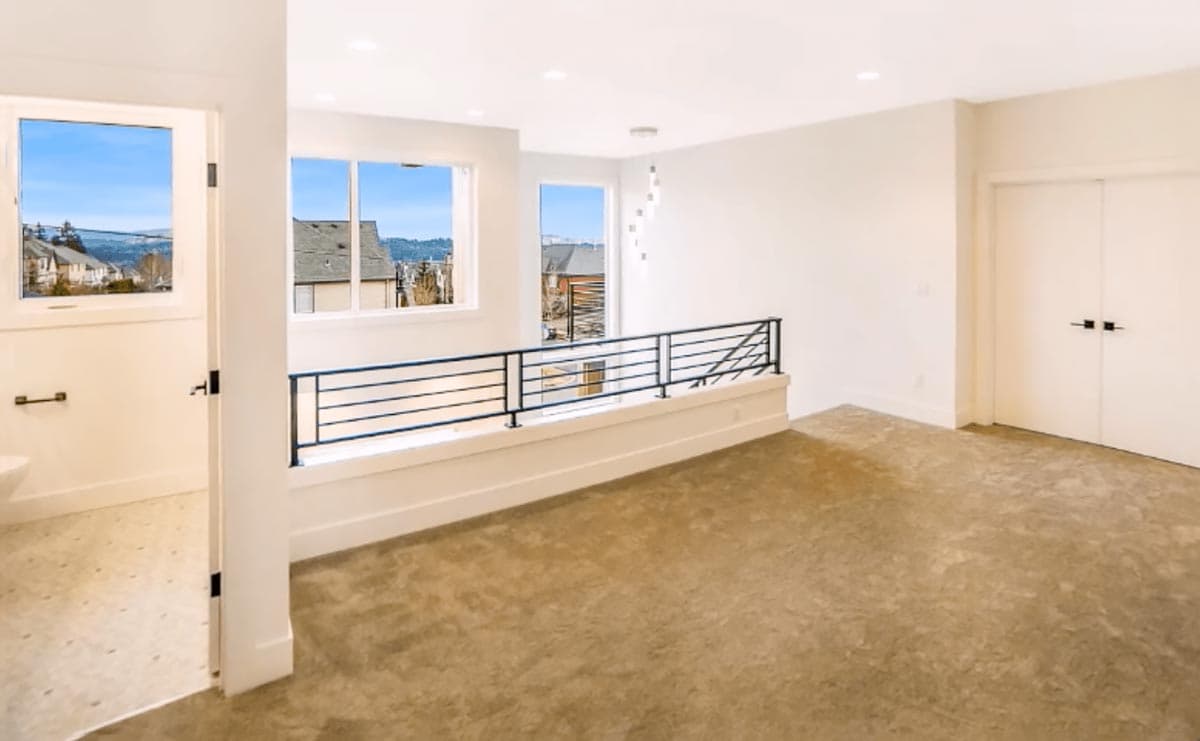 Interior view of a loft space with a metal railing, large windows showing exterior homes, and double doors.