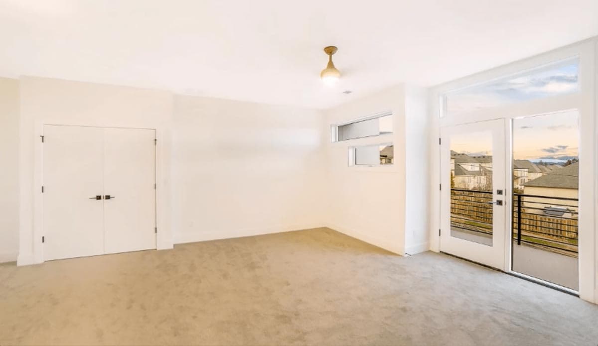 Interior view of a carpeted room with double doors, horizontal windows, and glass doors leading to a balcony with a view of houses.