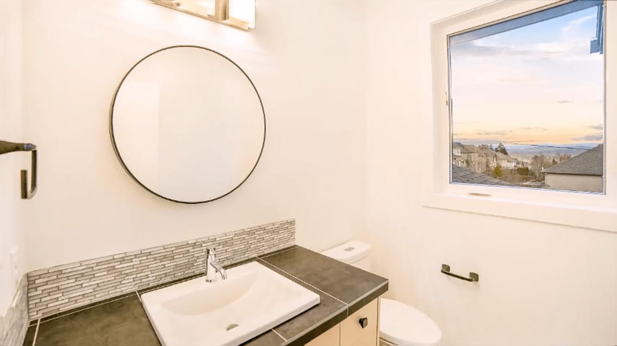 Powder room with vanity, circular mirror, mosaic tile backsplash, toilet, and large window with a view.