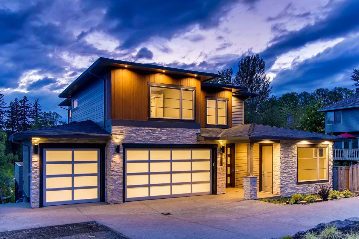 Two-story modern house plan exterior with a prominent gable roof, stone facade, wood siding, and two illuminated glass panel garage doors.