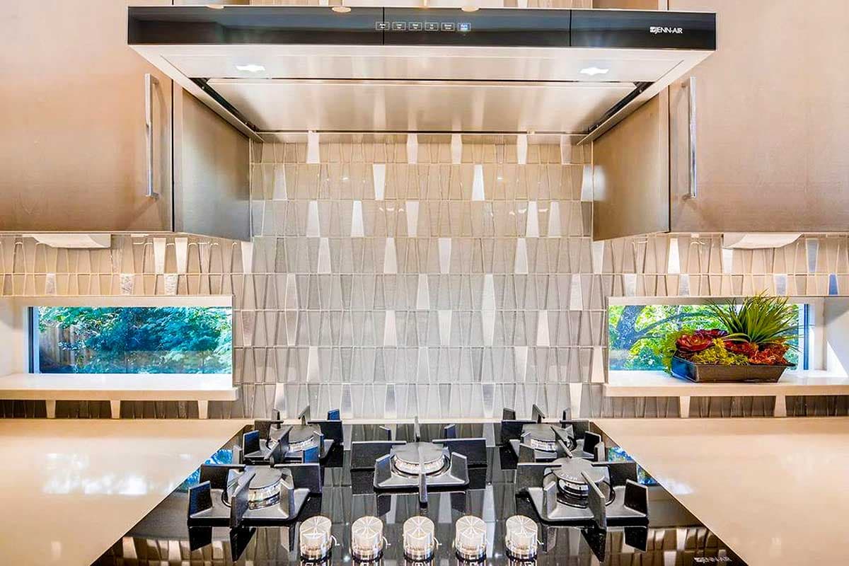 Kitchen features gas range, modern tile backsplash, Jenn-Air hood vent, and two small windows with greenery.