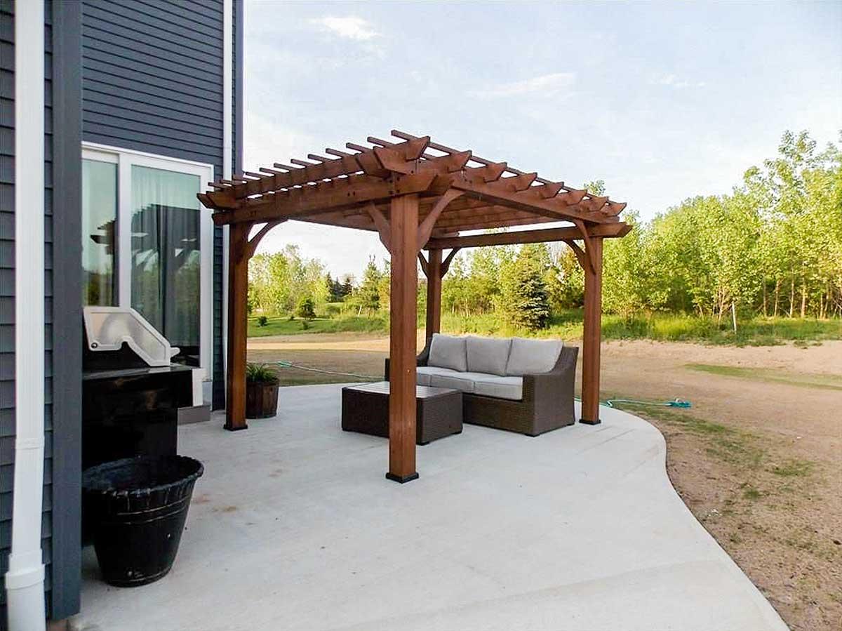 Outdoor patio with a wooden pergola, outdoor sofa, coffee table, and grill next to a house with dark siding.