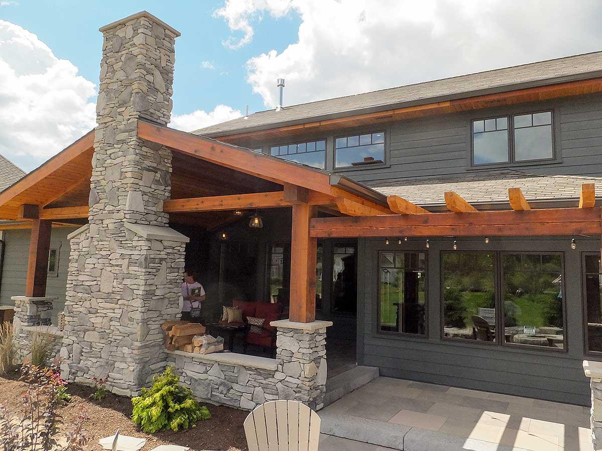 House plan exterior: Two-story home with a prominent stone chimney, exposed rafter tails, and a covered patio with a pergola.