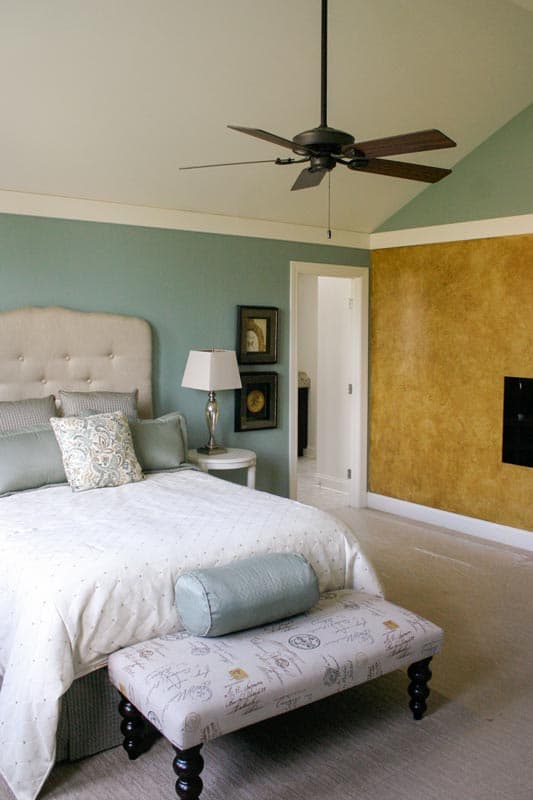 Bedroom with tufted headboard, decorative bench, ceiling fan, and textured accent wall. Doorway leads to en suite.