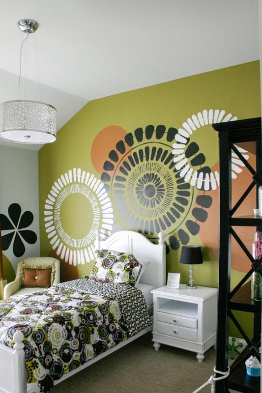 Bedroom with a white bed, patterned bedding, a lime green accent wall with large circular graphics, and a white bedside table.
