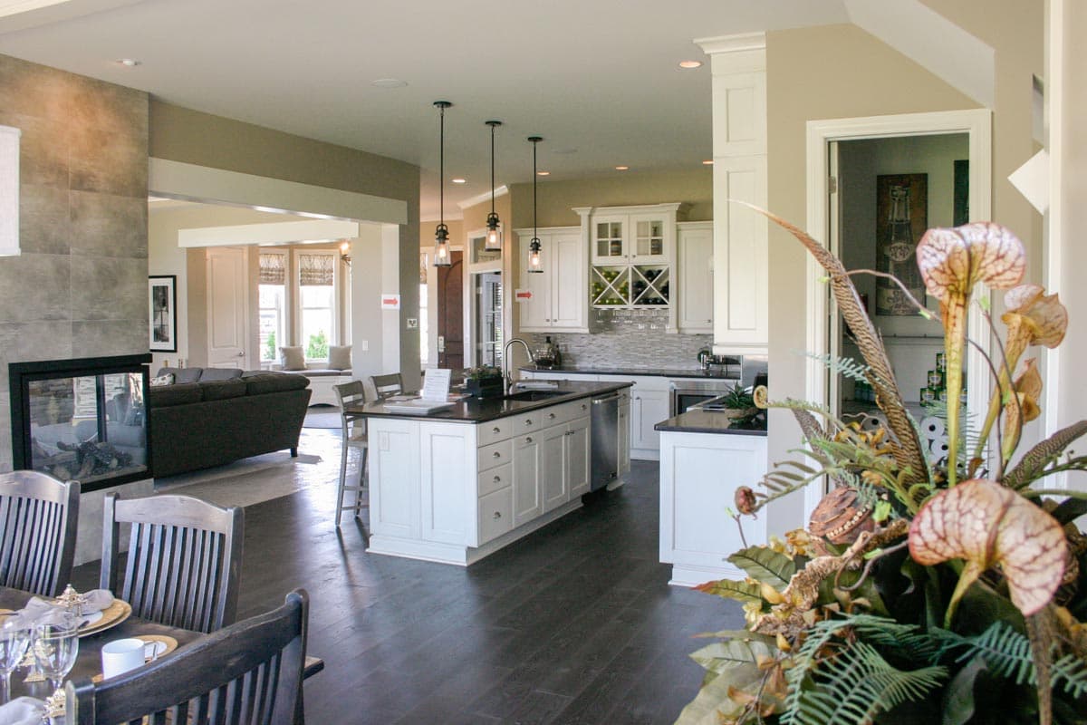 Interior view of a great room and kitchen with a stone fireplace, central island with sink, white cabinetry, and a seating area with bay windows.