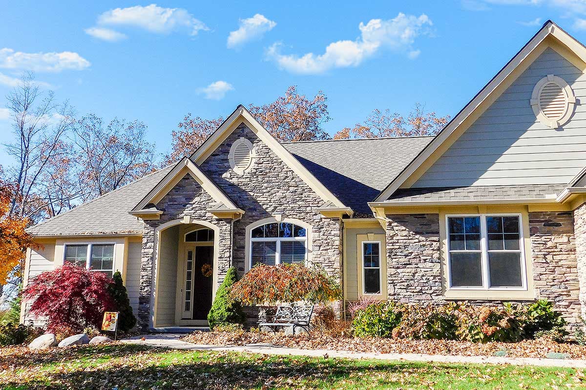 House plan exterior featuring stone accents, arched entry, gables with oval vents, and horizontal siding.