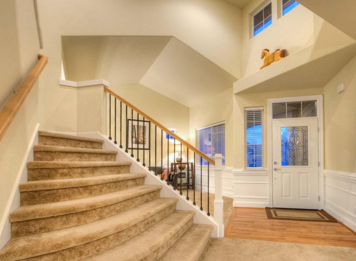 Interior view of a house entrance with carpeted stairs, wrought iron banister, decorative glass front door, and two windows.