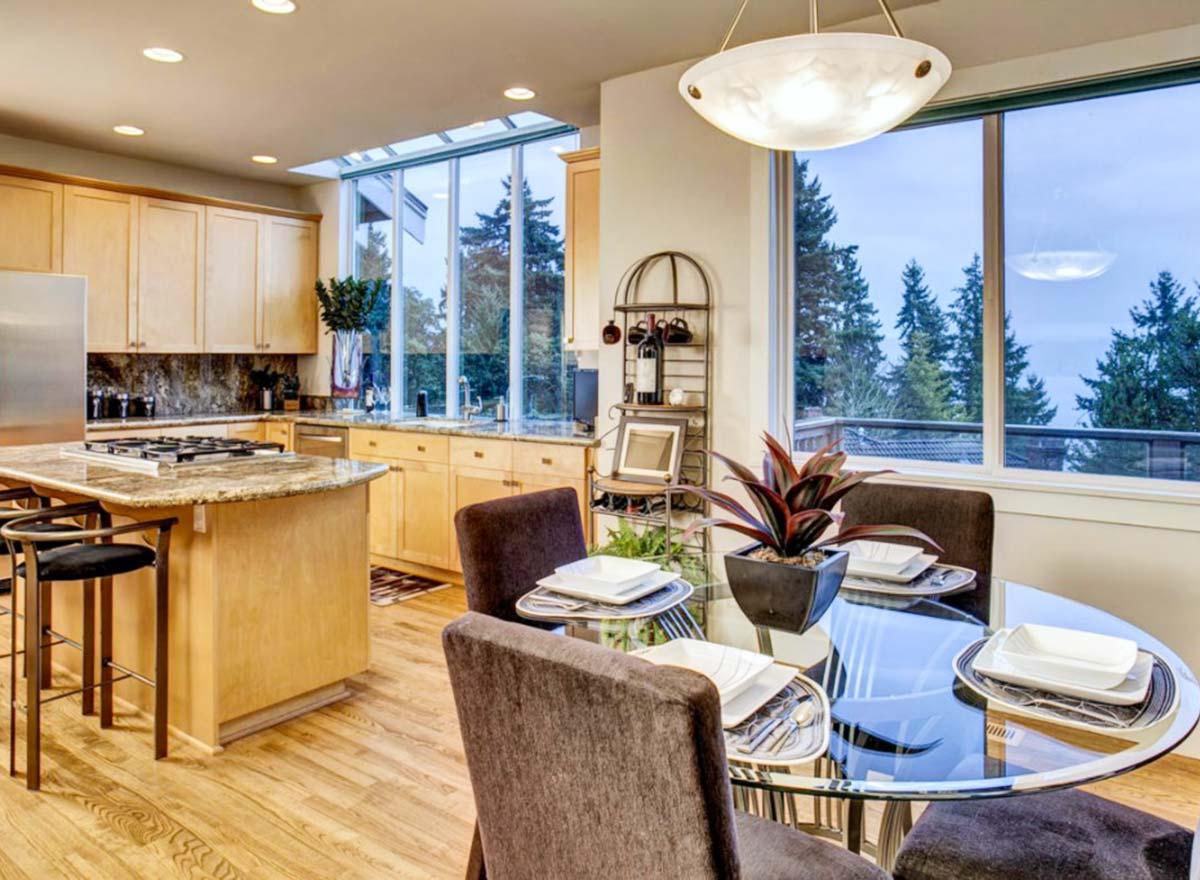 Modern kitchen with island, gas range, and dining area. Large windows and skylight offer natural light and forest views.