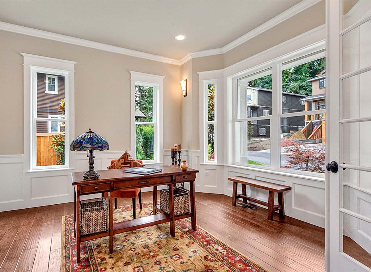 Interior view of a home office with a desk, bench, and large windows overlooking a street with houses.