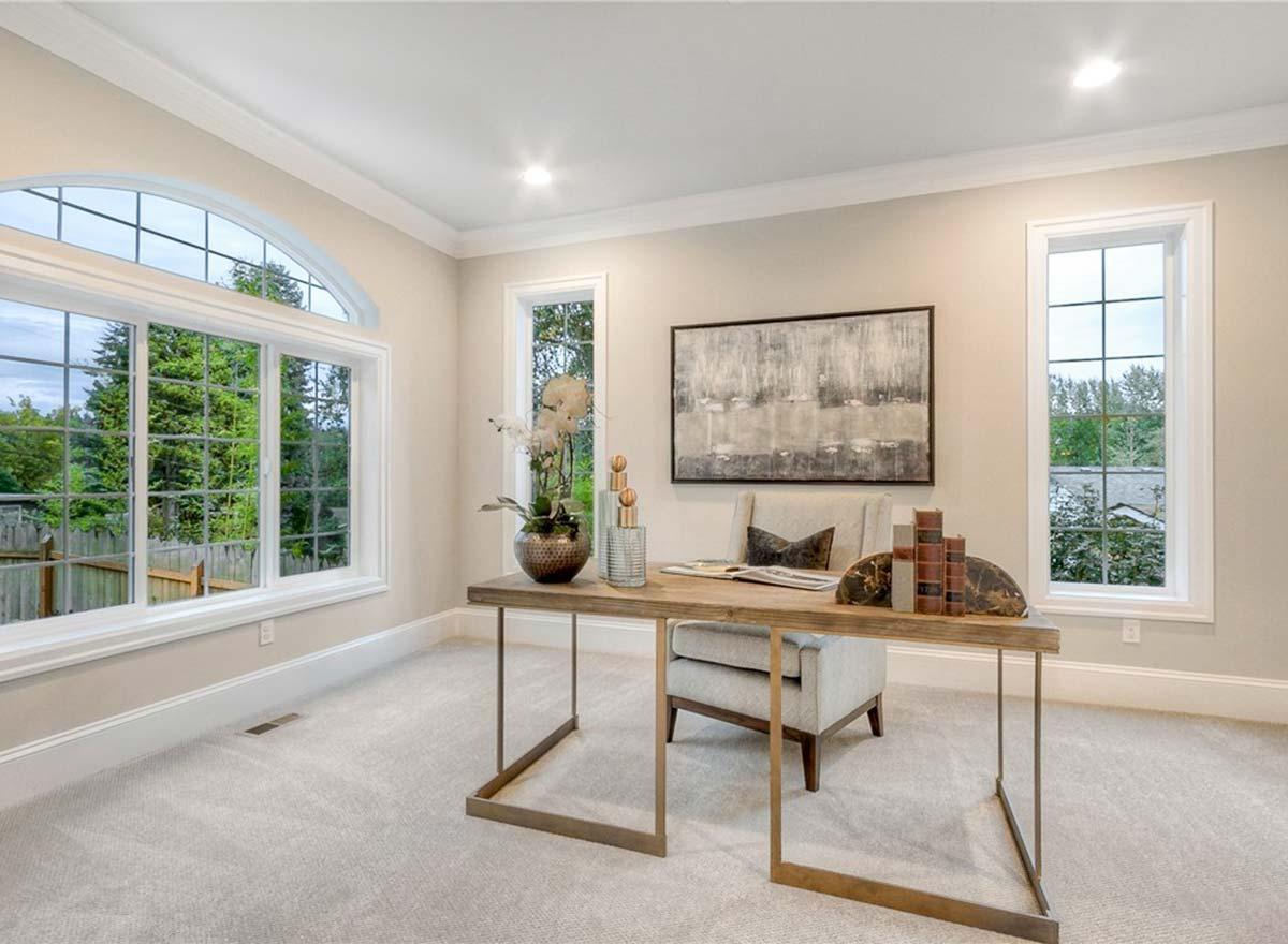 Home office with a wooden desk, chair, large arched window, and two rectangular windows. Neutral carpet and walls.