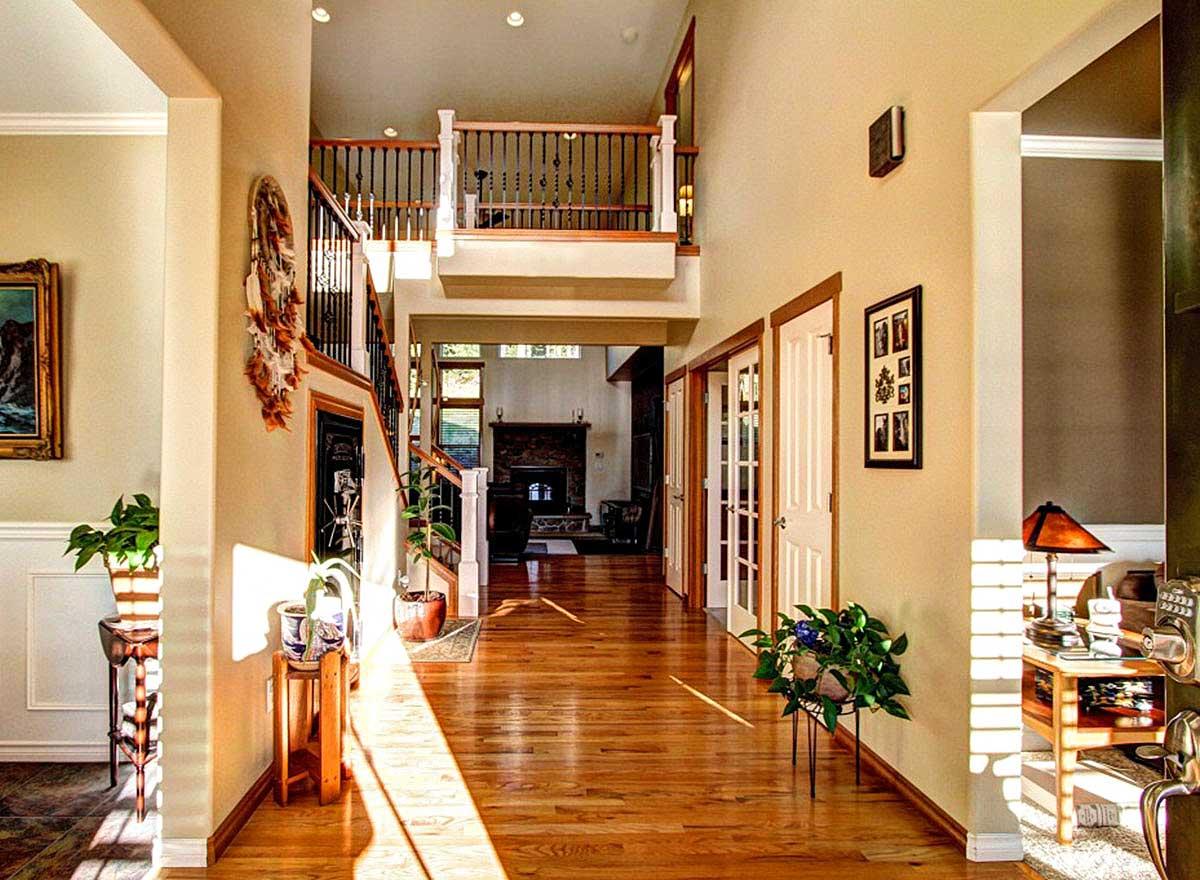 Interior view of a house with a grand staircase, second-floor landing with black spindles, and a view into a living room with a fireplace.