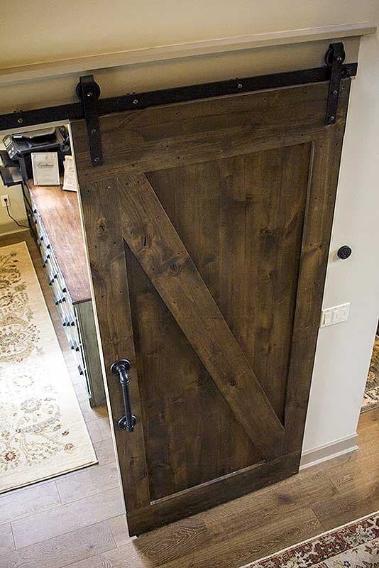 Rustic wood barn door with black hardware and a diagonal plank design. Visible in a hallway with a patterned rug.
