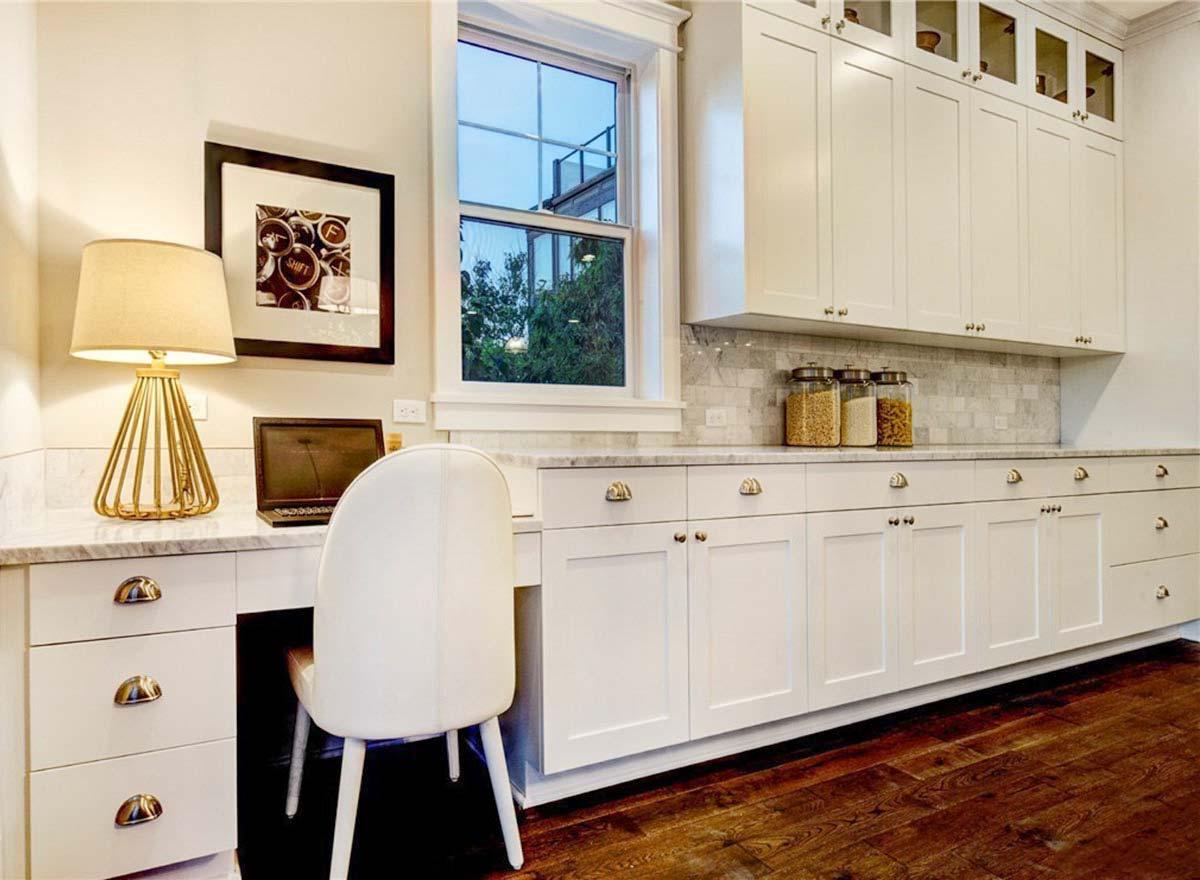 Built-in desk with drawers and upper cabinets with glass fronts, adjacent to a window and white cabinets.