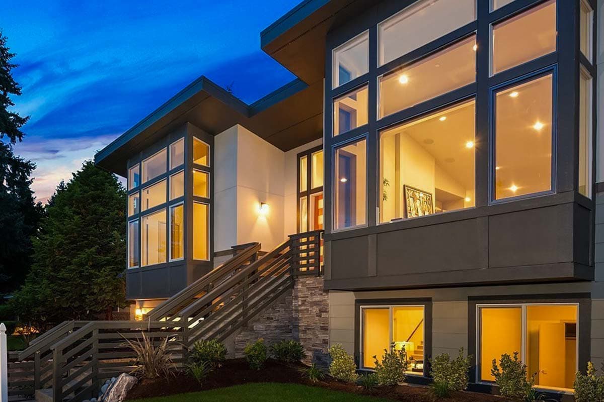 Modern house exterior with multiple stories, large glass windows, a cantilevered section, and a stone-clad base with a wooden staircase.