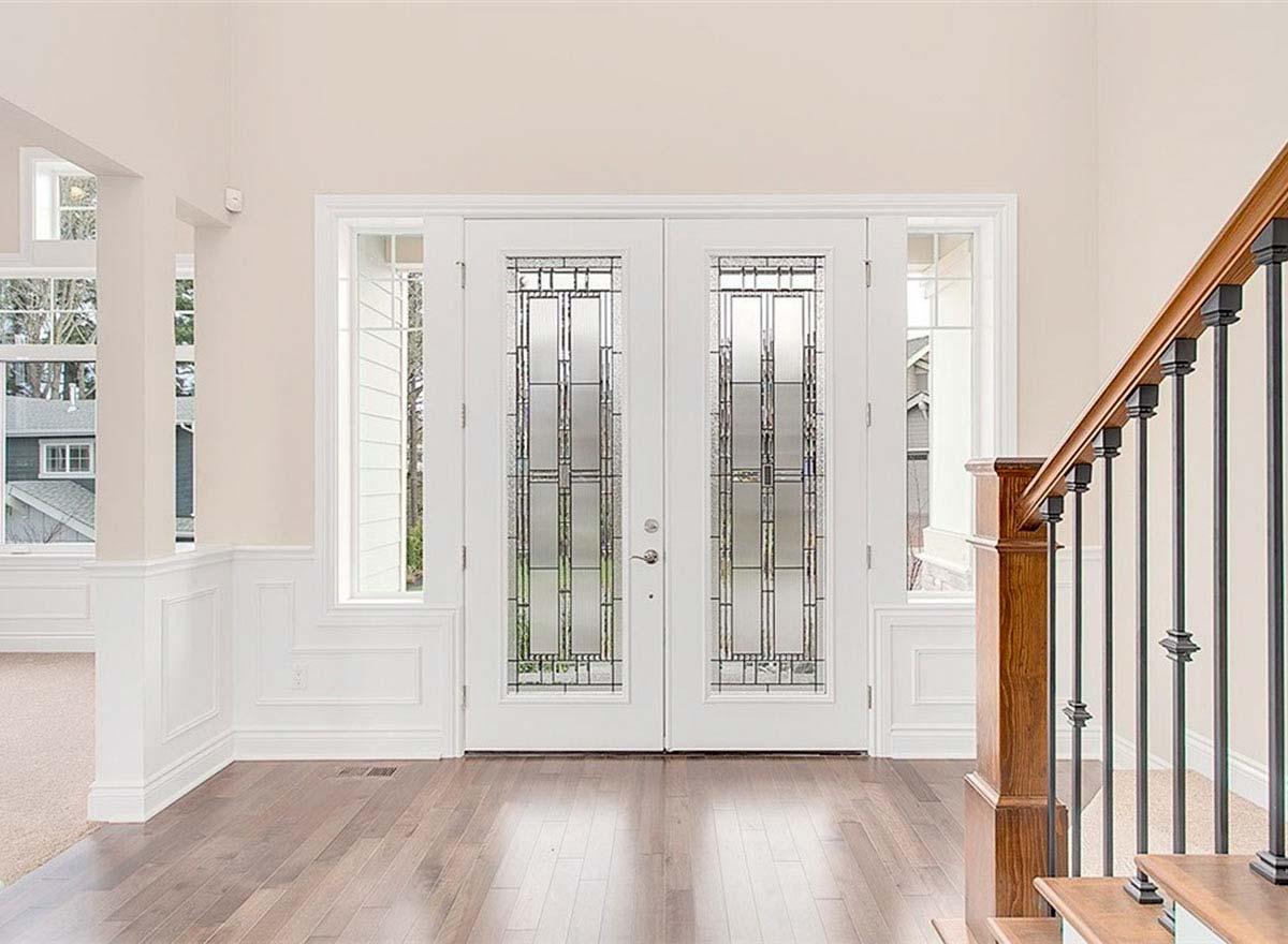 Interior view of a foyer with double glass-paneled doors, sidelights, wainscoting, and a wooden staircase with metal spindles.