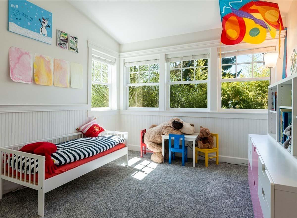 Children's bedroom with white wainscoting, a twin bed with striped bedding, and multiple windows overlooking trees.