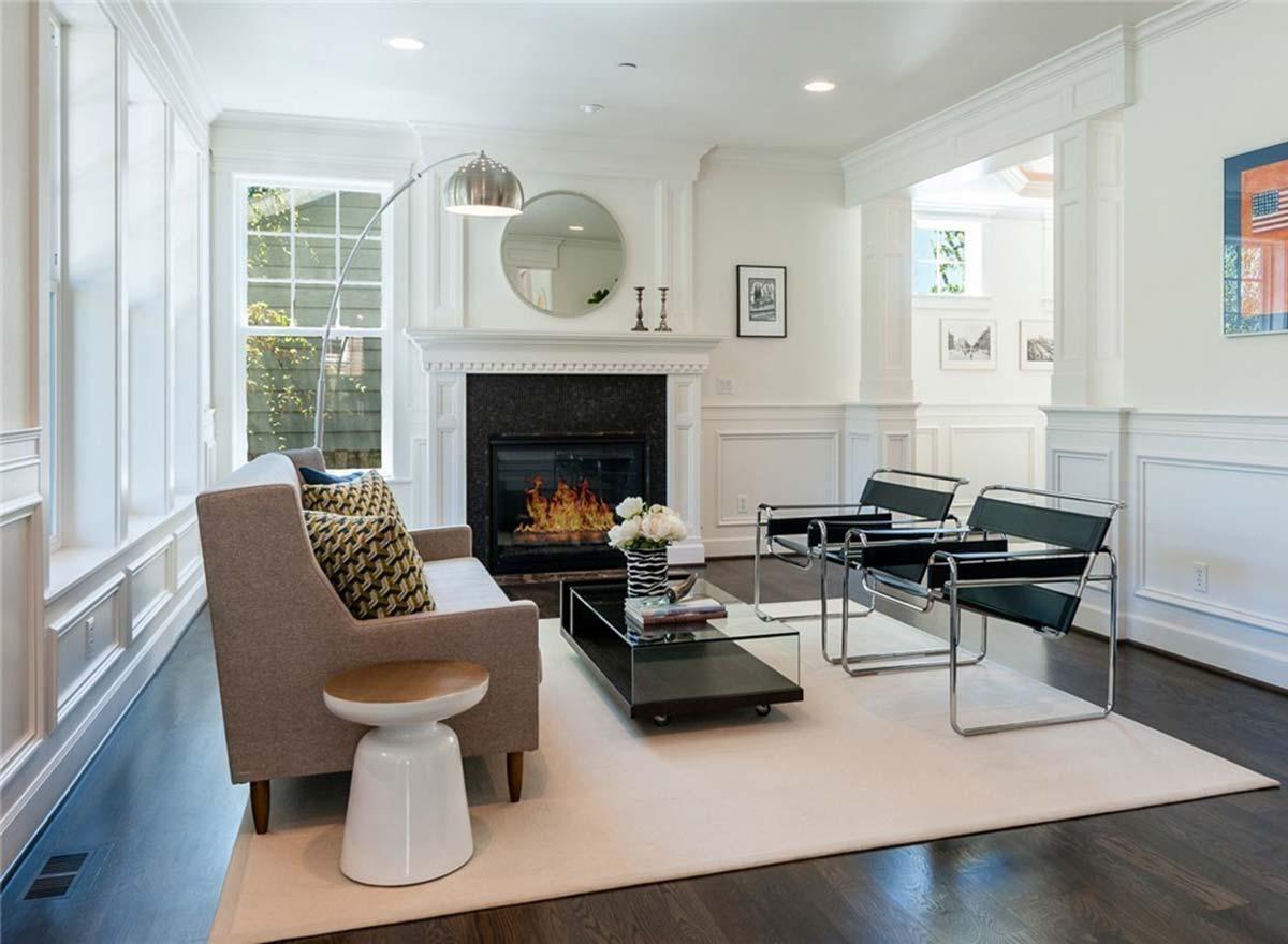Interior view of a living room with a fireplace, wainscoting, large windows, and modern furniture.