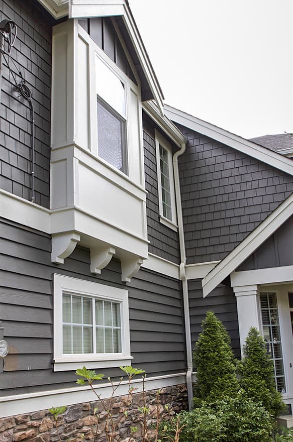 House plan exterior shows a dark gray shingle-clad facade with a projecting bay window and stone foundation.