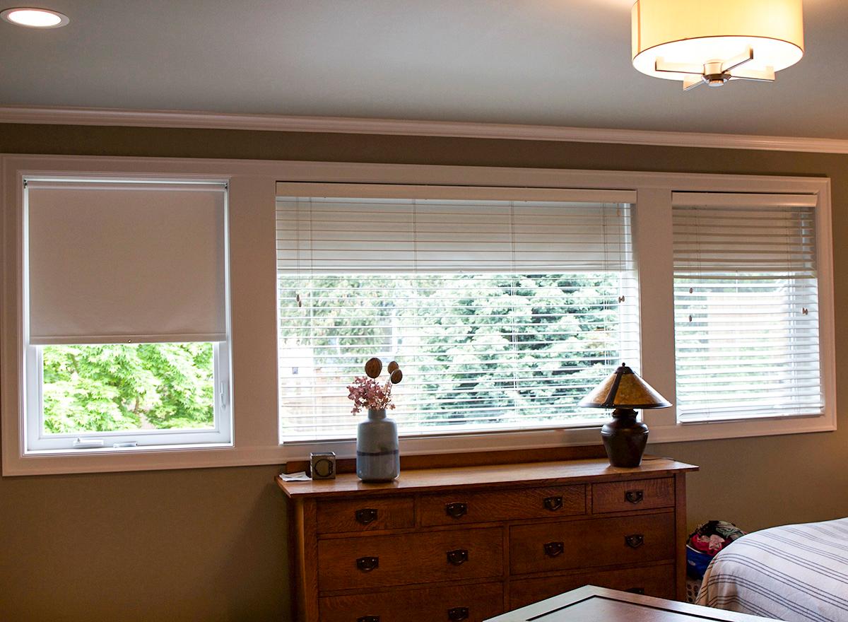Bedroom with three windows: one roller shade, two with blinds. Wooden dresser and lamp visible.