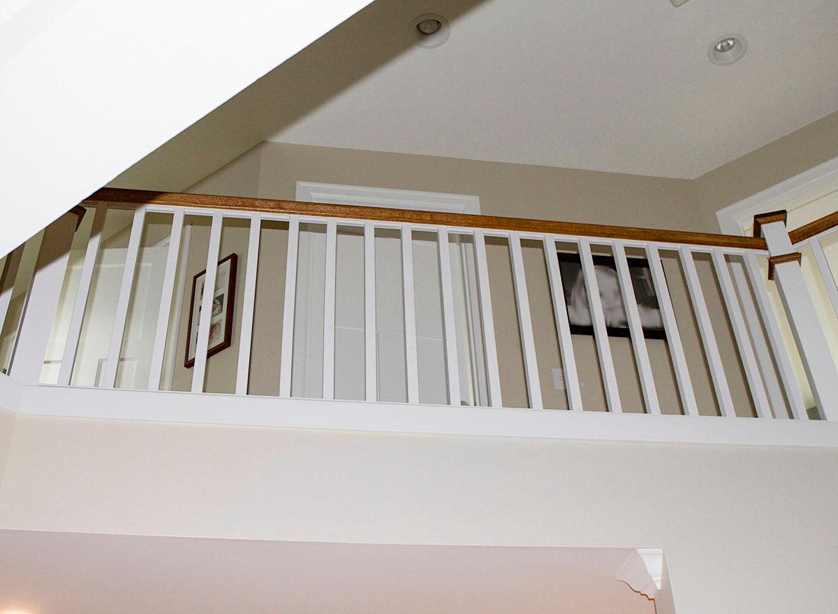 Interior view of a hallway overlook with white balusters and wood handrail, featuring two doors and framed artwork on the wall.